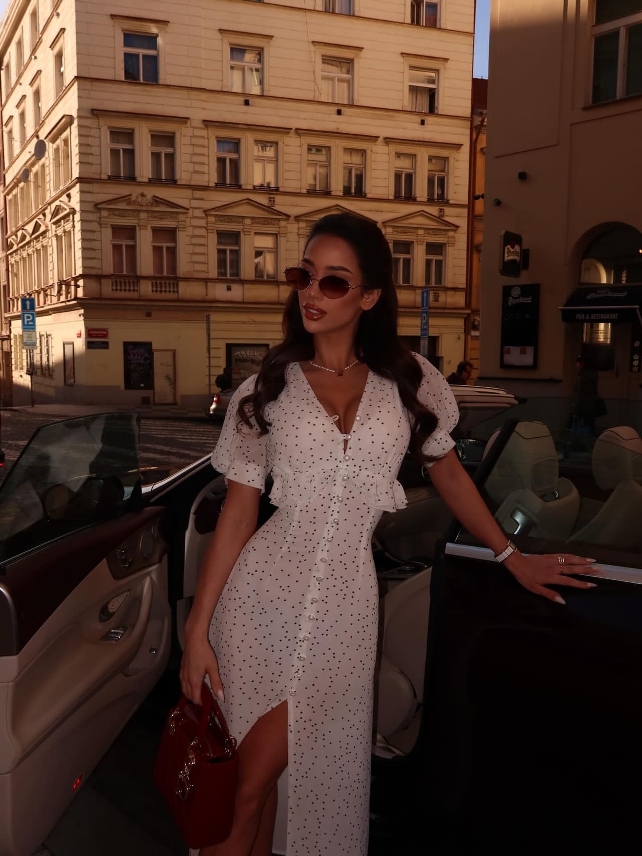 A woman in a white polka-dot midi dress with sunglasses stands by an open car door on a city street, holding a red bag.